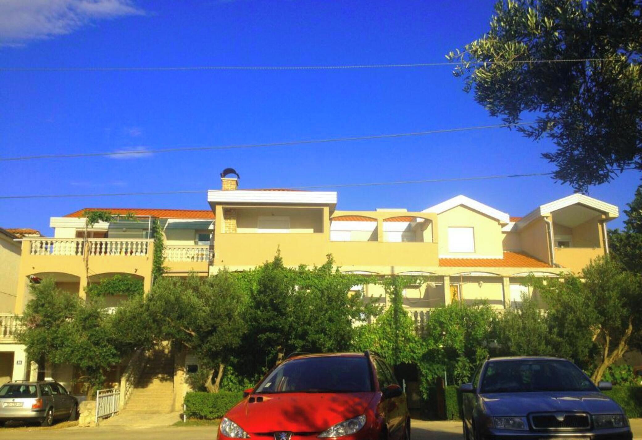 Un immeuble d’habitation doté de nombreux balcons, une voiture rouge garée devant, et un ciel bleu dégagé.