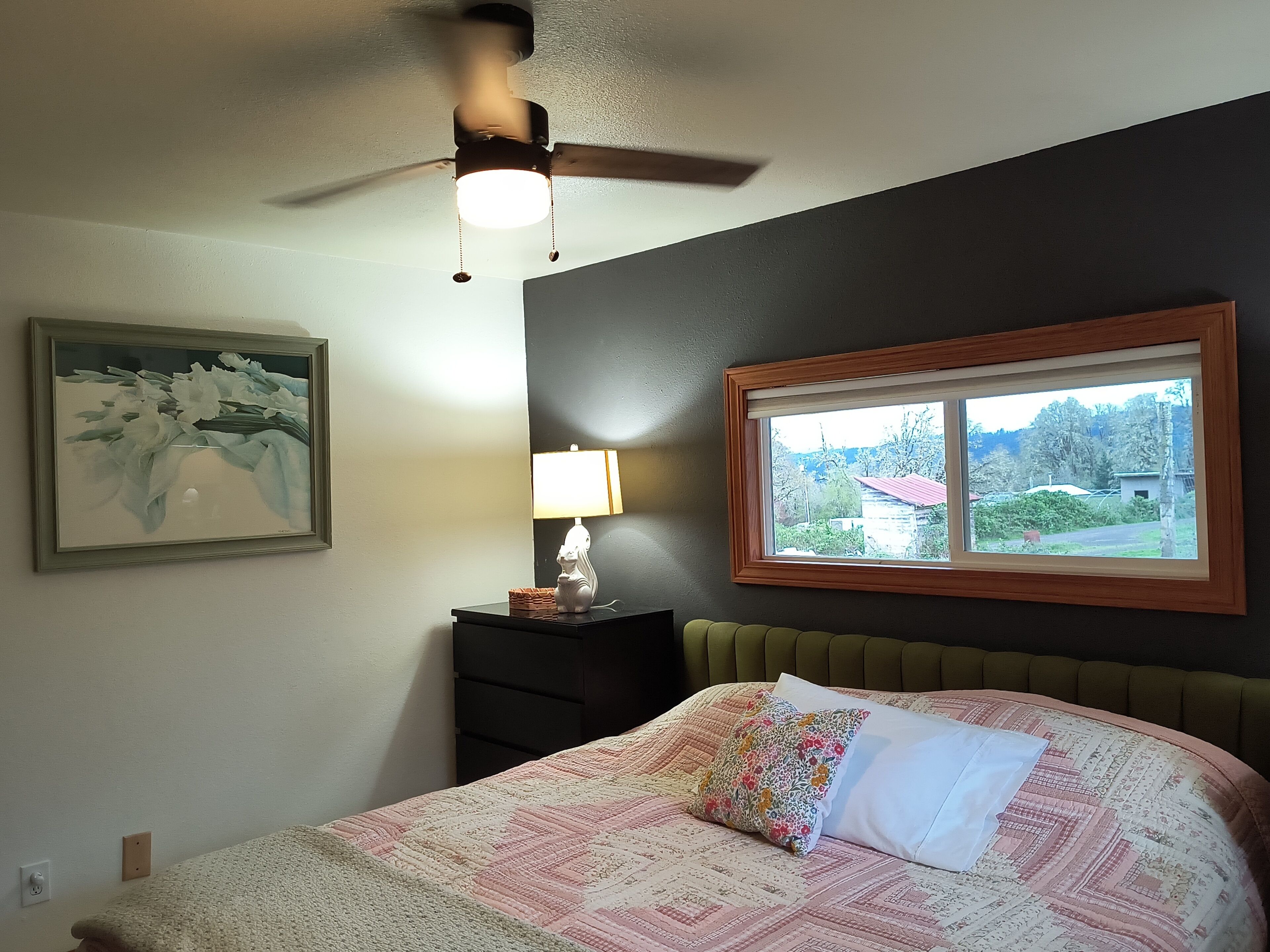 Une chambre à coucher comprenant un lit, une table de chevet, une lampe, un ventilateur de plafond, un tableau encadré et une fenêtre donnant sur un beau paysage.