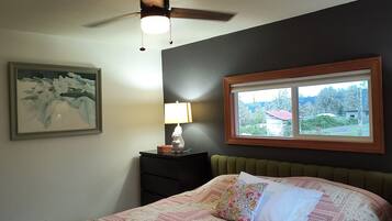Une chambre à coucher comprenant un lit, une table de chevet, une lampe, un ventilateur de plafond, un tableau encadré et une fenêtre donnant sur un beau paysage.