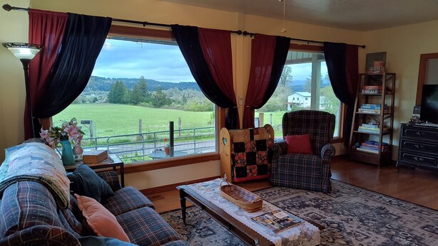 A living room with a sofa, armchair, and a table with magazines. There is a view of a green field through large windows. - Charming 2-bedroom farmhouse in Willamina's relaxing countryside. (Willamina)