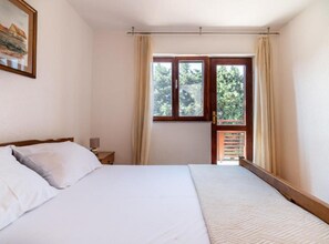 Quarto com cama, mesa de cabeceira, janela com cortinas e vista para o verde lá fora.