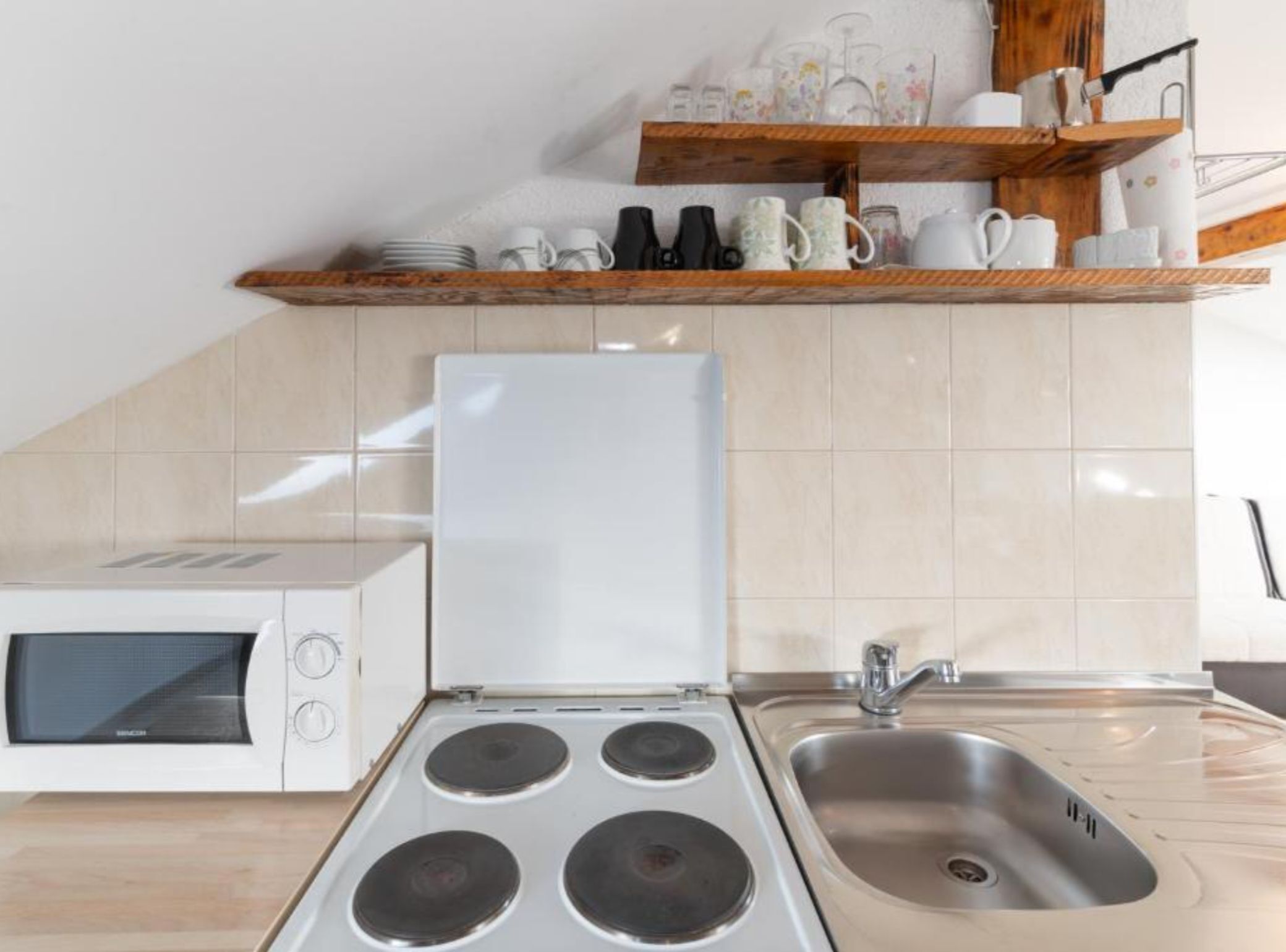 A kitchen with a microwave, stove, sink, and a shelf with dishes and cups.