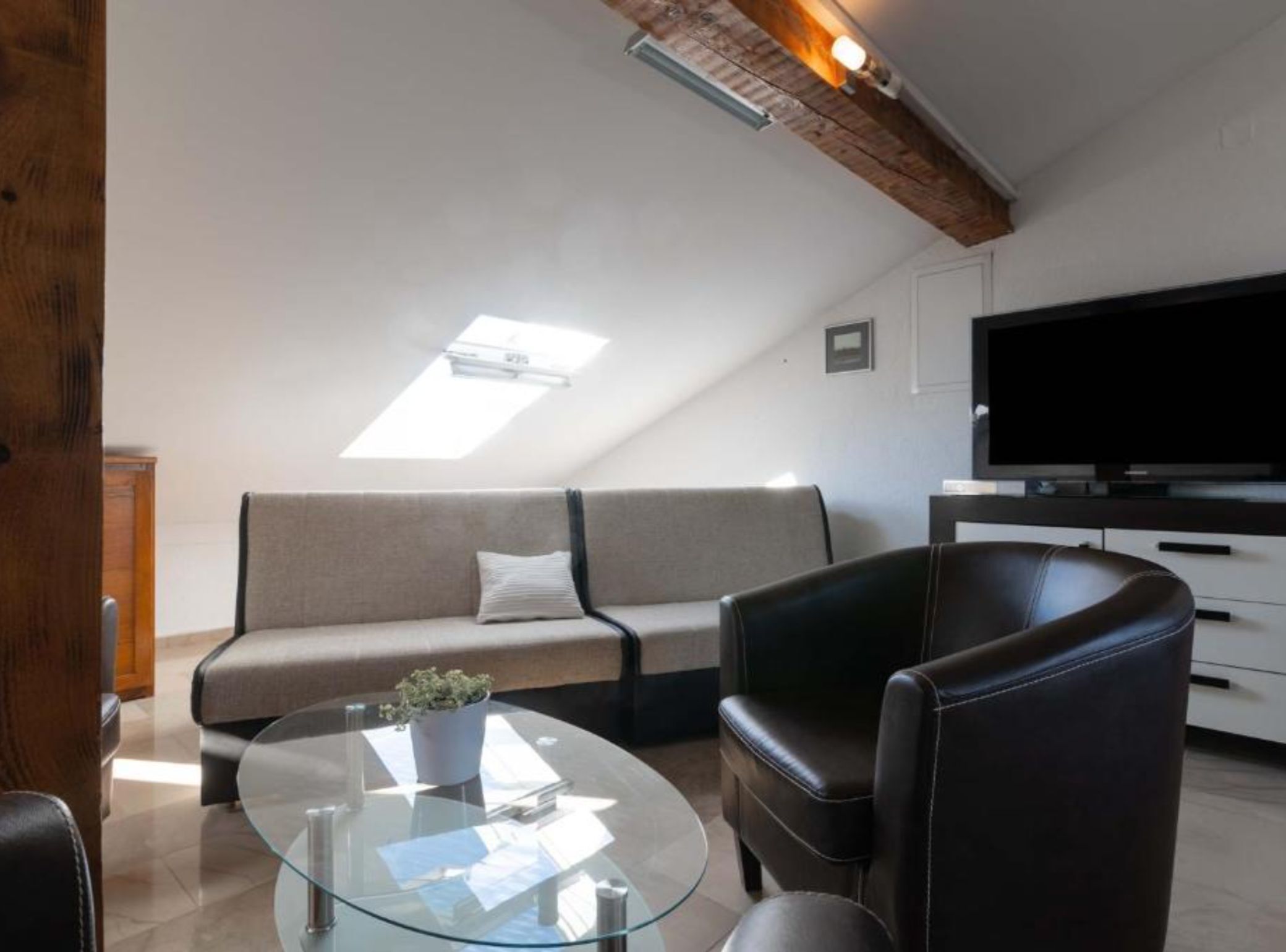 A modern living room with a glass coffee table, a sofa, a leather armchair, a television, and a skylight.