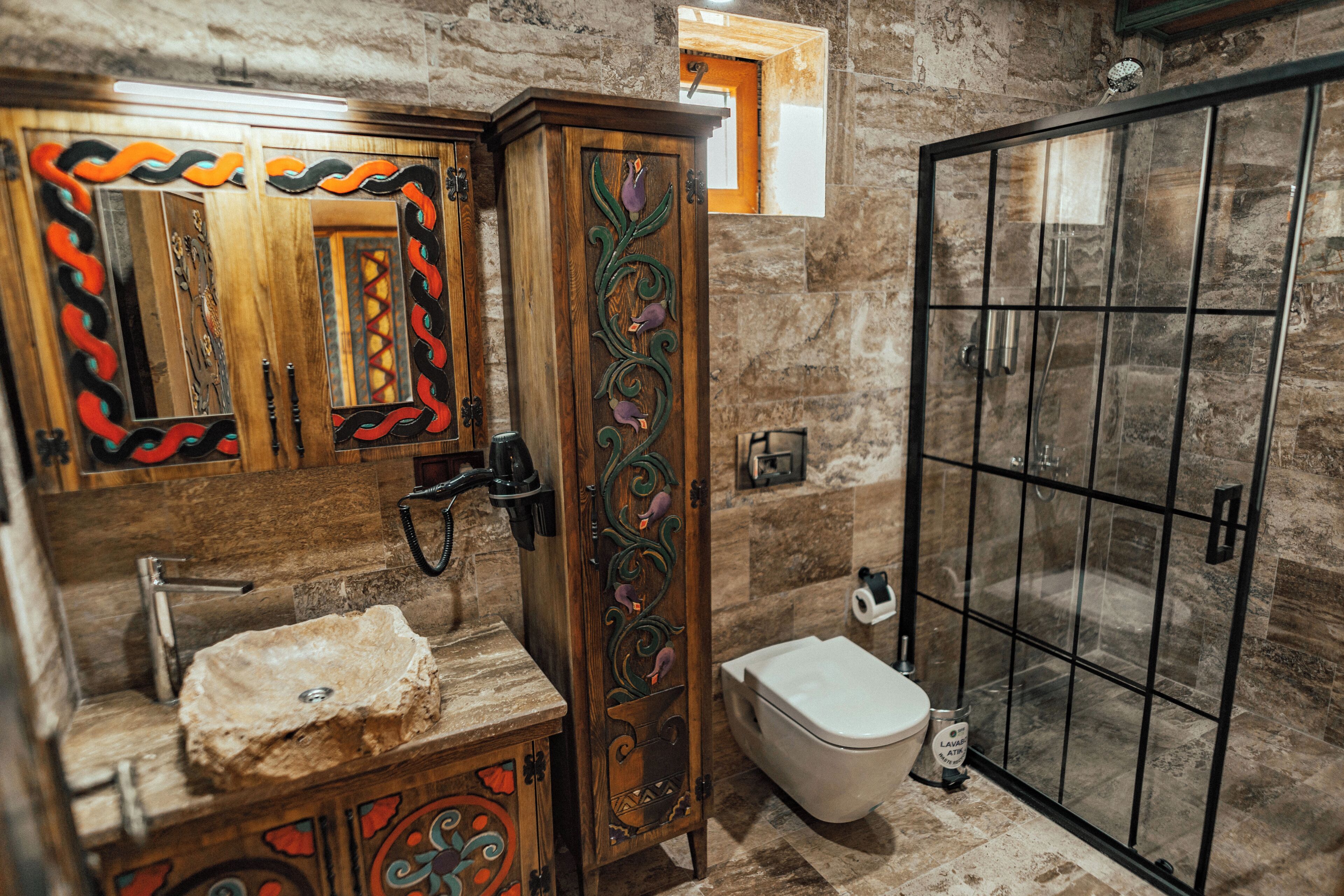 A bathroom with a stone sink, wooden cabinet with intricate carvings, a toilet, and a glass-enclosed shower.