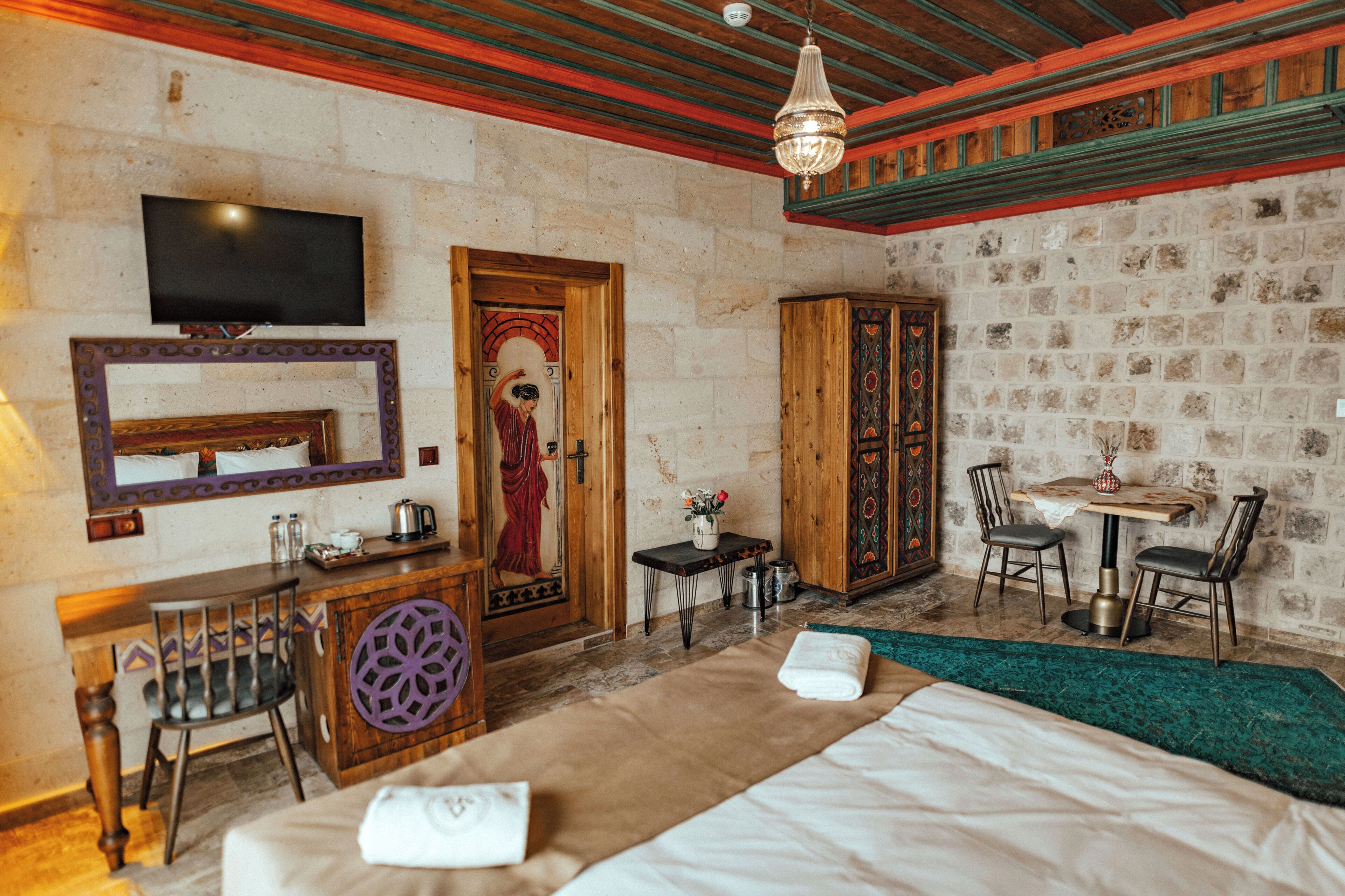 A hotel room with a bed, a desk, a television, a mirror, a door with a traditional design, and a small table with chairs.