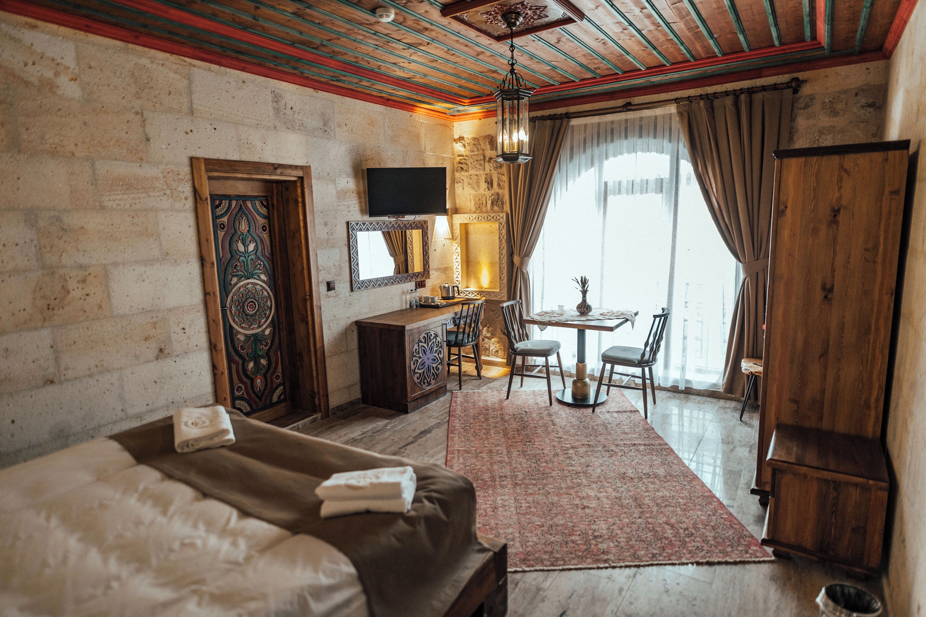 Habitación de hotel con cama, escritorio, silla, televisor y un gran ventanal con cortinas.