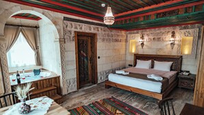 A room with a bed, a small table with a vase, a window with curtains, a wooden door, and a stone wall. - Kallista Cappadocia (Nevsehir)