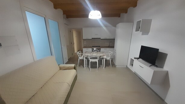A living room with a sofa, a dining area with chairs, a kitchen with cabinets, and a television on a stand. - Vacation home La Cilinella del.Salento (Copertino)