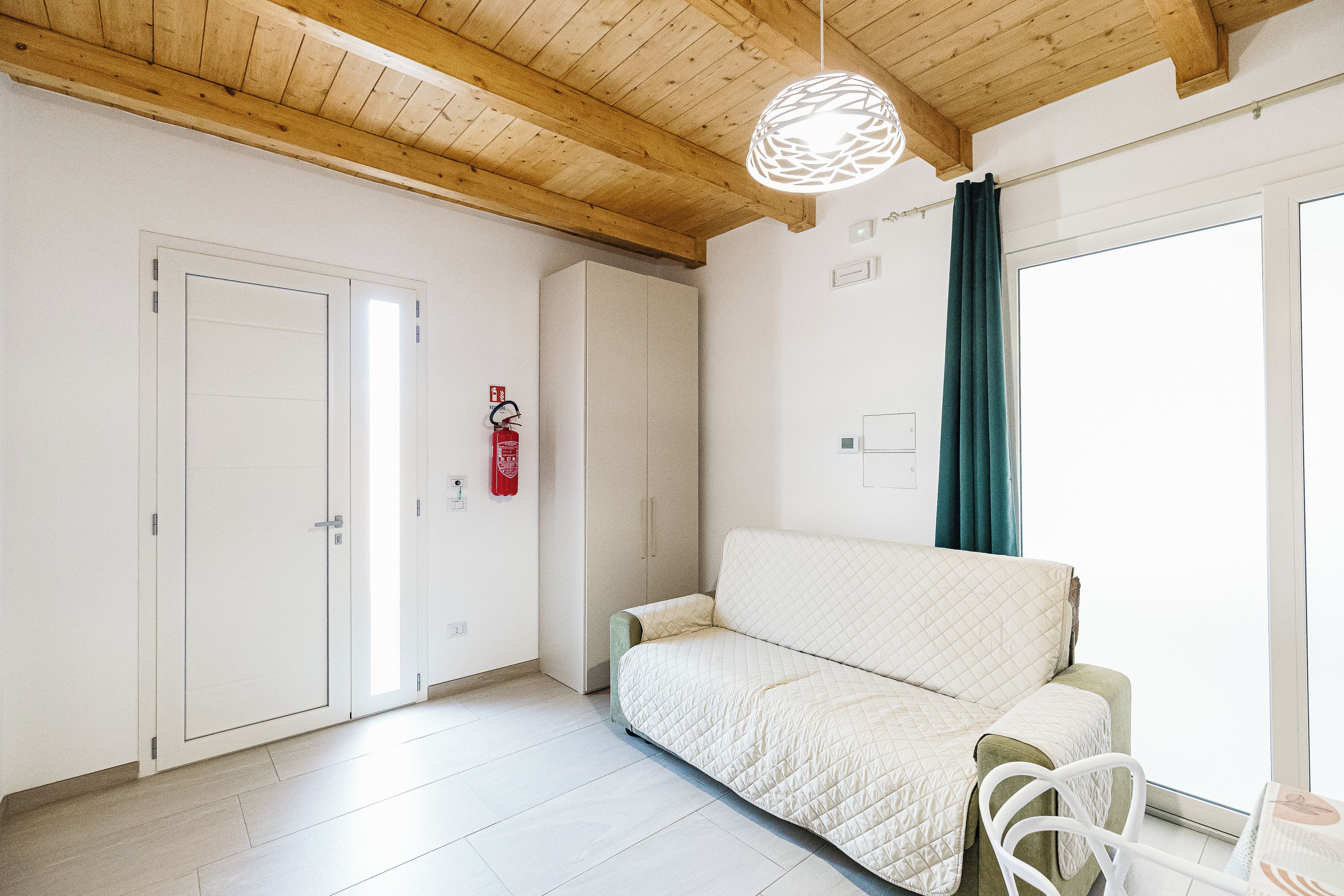 Ein Zimmer mit einer weißen Couch, einer Holzbalkendecke, einer weißen Tür und einem Fenster mit Vorhängen.