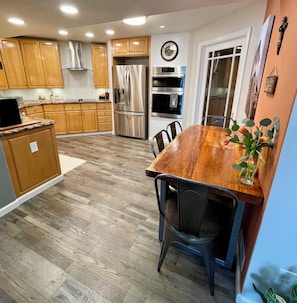 Une cuisine avec des meubles en bois, des appareils électroménagers en acier inoxydable, une table à manger avec des chaises et une horloge au mur.