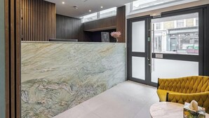 A reception area with a marble counter, a yellow upholstered chair, and a view of the street through a glass door. - Journeys Kings Cross - Hostel (London)