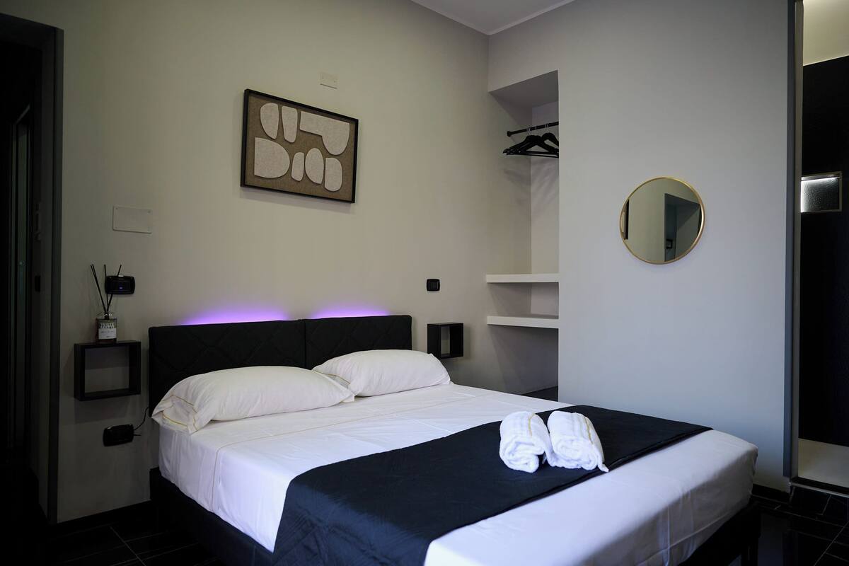 Una cama bien hecha con sábanas blancas, cabecero y colcha negros. En la pared, un espejo redondo, una obra de arte abstracta enmarcada y una mesita de noche con una lámpara.
