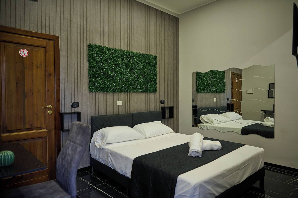 Habitación de hotel con cama, una silla, puerta de madera, panel de pared verde, espejo y estante pequeño.