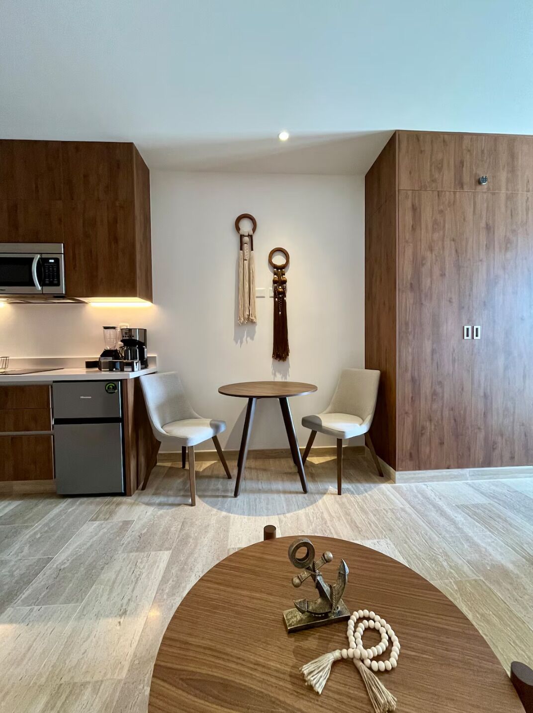 Een moderne keuken met een magnetron, koffieapparaat en een kleine koelkast. Er staat een ronde houten tafel met stoelen en een decoratief object erop.