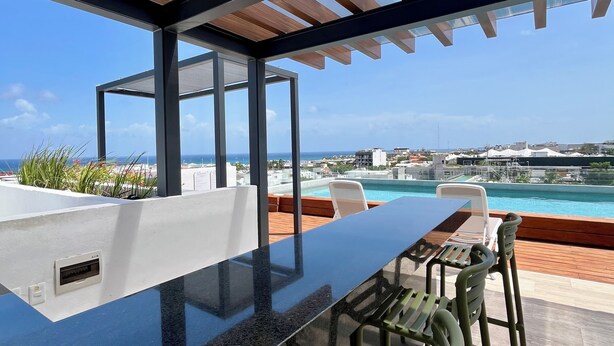 Uma área de refeições moderna ao ar livre, com estrutura de vidro, piscina e vista para o oceano e a cidade.