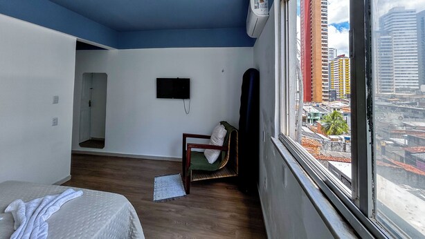 A room with a bed, a chair, a TV, and a window with a city view. - Modern loft in Belém with solar energy - At Doca Umarizal - downtown (Belém)