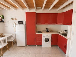A compact kitchen with red cabinets, a white refrigerator, a microwave, a washing machine, and a dining area with a white table and chairs.
