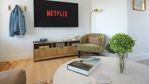 Olohuoneessa on litteänäyttöinen televisio, jossa näkyy Netflix, vihreä nojatuoli, kirjoja sisältävä sohvapöytä, lattiavalaisin ja vihreä huonekasvi.