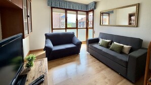 A living room with a TV, two sofas, a wooden coffee table, and a large window with a view of buildings. - Las Amapolas Apartment - 12018 (Ajo)