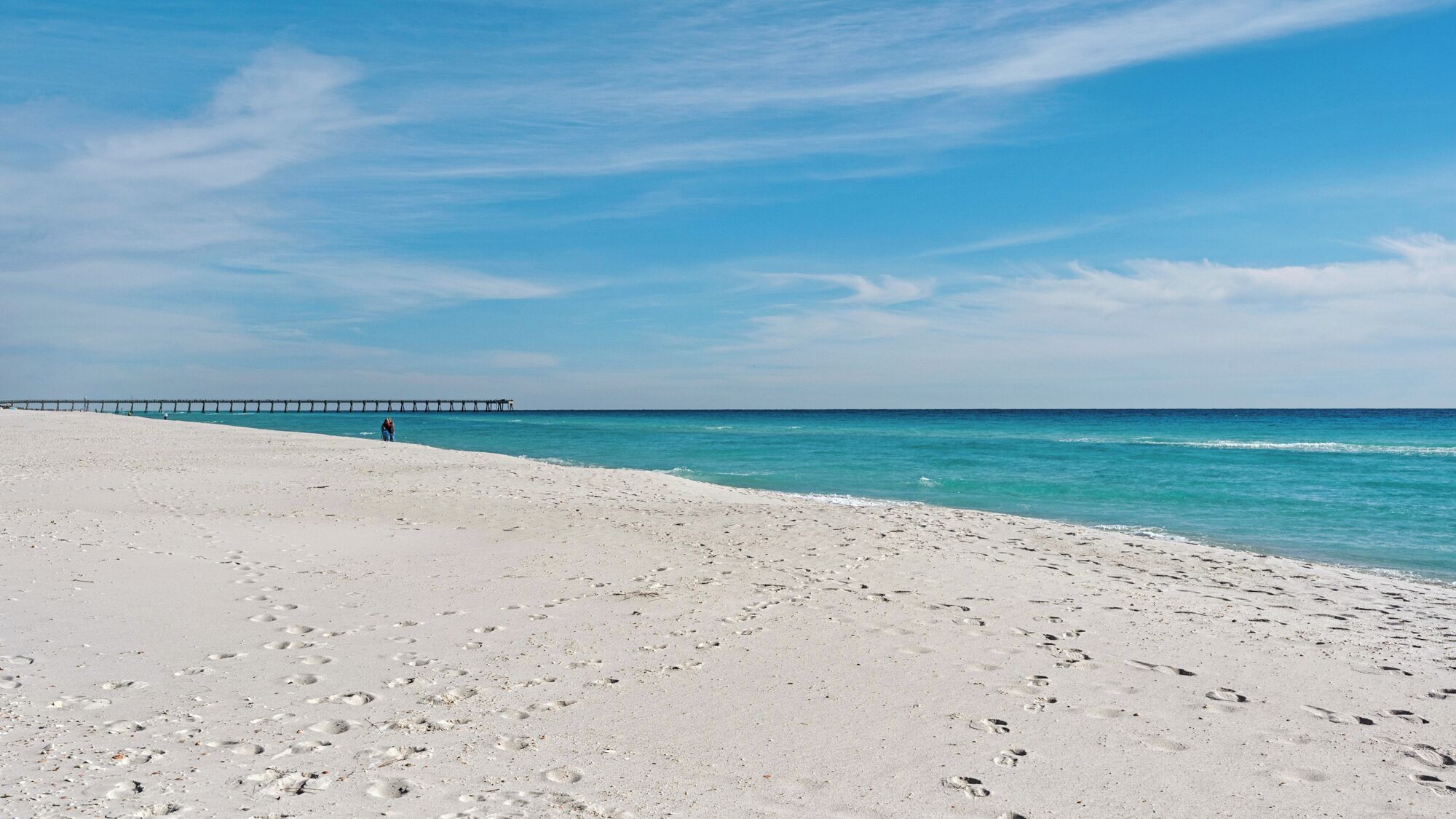 A beach with white sand and a pier in the distance.