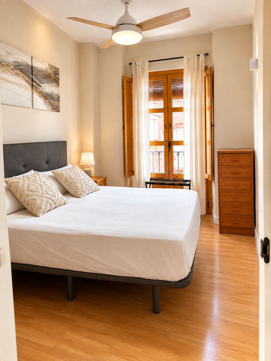Une chambre à coucher comprenant un lit, un ventilateur de plafond, une table de chevet, une commode et une fenêtre avec des rideaux.