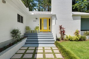 A white building with a yellow door, numbered 3514, and a small porch with two chairs. - Modern Home Close to UF. Pet-Friendly. EV Charger. Patio and BBQ (Gainesville)