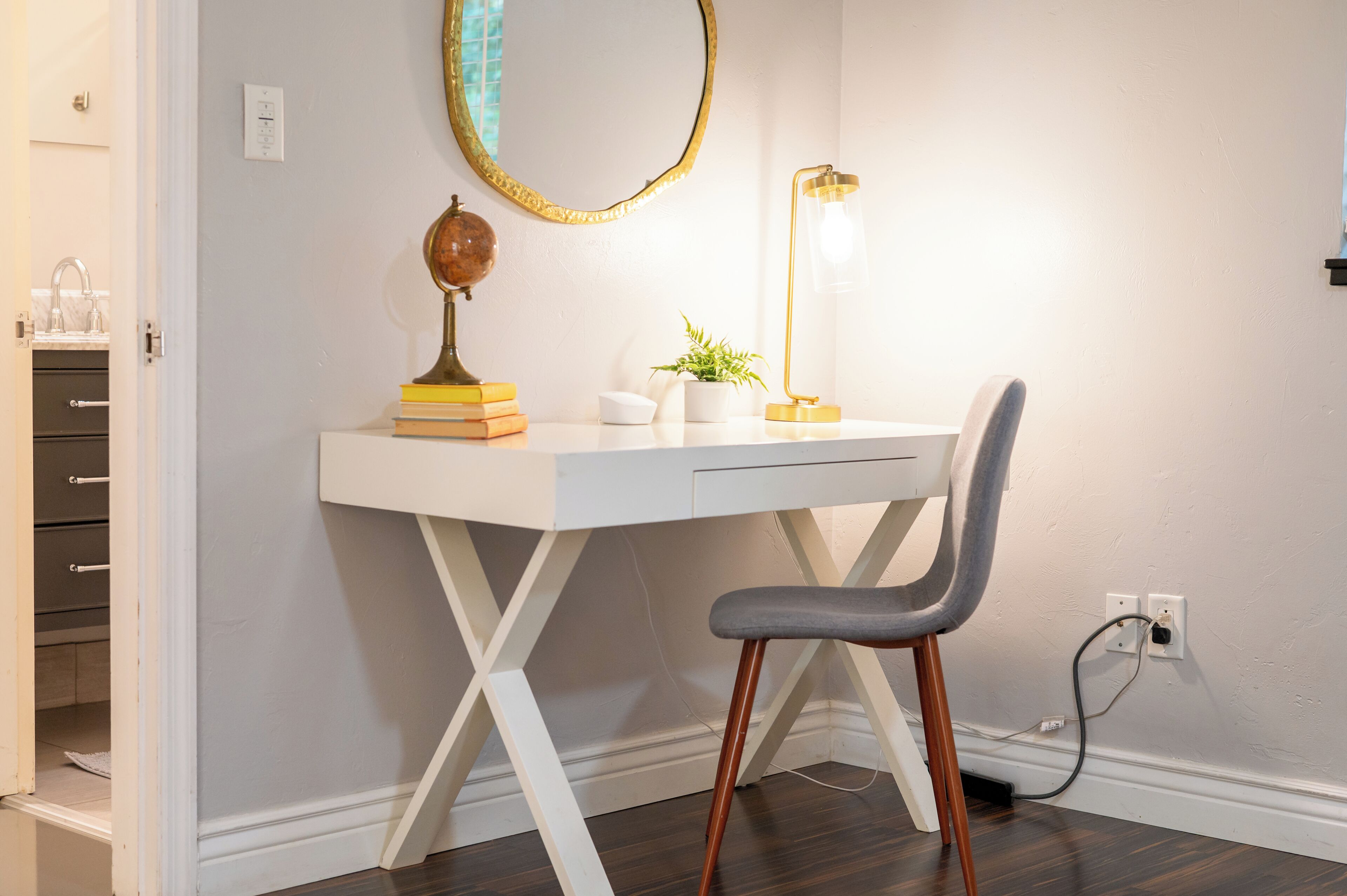A white desk with a globe, books, and a plant, a gray chair, and a gold-framed mirror on a wall.