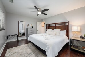 Een slaapkamer met een bed, een plafondventilator, een nachtkastje met een lamp, een dressoir, een spiegel en een vloerkleed.