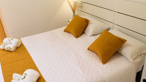 Una cama bien hecha con almohadas blancas y mostaza, una colcha blanca y un par de toallas enrolladas en el suelo.