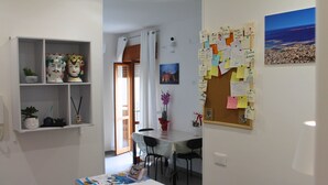 Una habitación con una mesa de comedor, sillas, un tablón de anuncios con notas, una planta en maceta y una estantería con adornos.