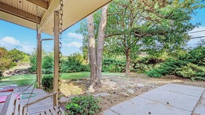 Une terrasse couverte avec un banc en bois, un chemin de pierre et une vue sur un jardin aménagé, avec des arbres et une pelouse.