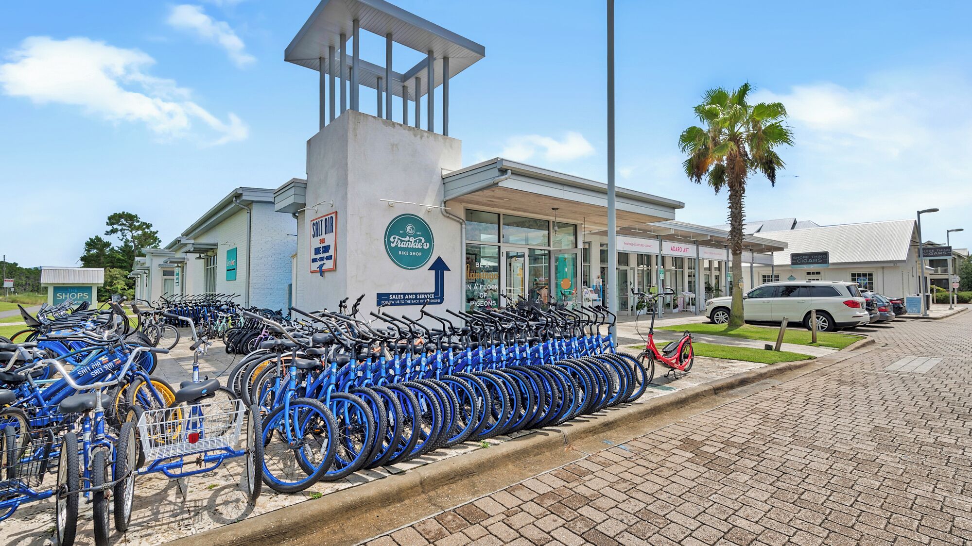 A bike rental station with multiple bicycles lined up in front of a building with a sign that reads "Frankie's Bike Shop".