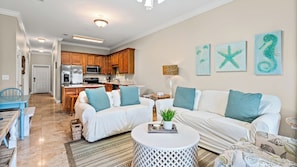 A spacious living room with a white sofa set, a dining table, and a kitchen in the background.