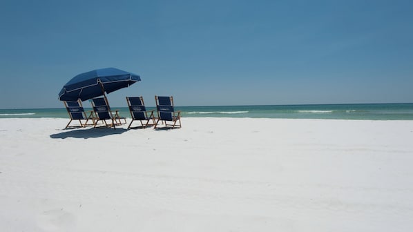 A beach with three lounge chairs under a blue umbrella, facing the ocean.