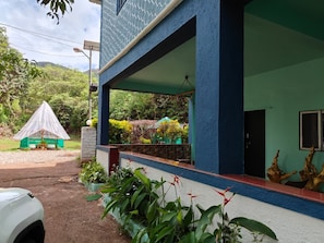 A building with a covered porch, a small structure with a conical roof, and lush greenery.