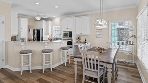 A modern kitchen with white cabinetry, a central island with stools, a dining area with a wooden table and chairs, and a hanging light fixture. - 4 Bedroom 3 Bath walking distance to everything + hot tub! (Holden Beach)