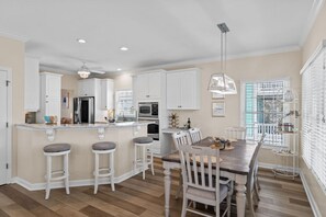 Una cocina moderna con armarios blancos, una isla central con taburetes, un comedor con mesa y sillas de madera, y una lámpara colgante.