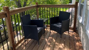Una terraza de madera con dos sillas de mimbre y una mesa de vidrio, rodeada por una barandilla de madera.