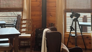 Interior de una cabaña de madera con una chimenea central, una mesa de comedor con sillas y una cámara sobre un trípode.