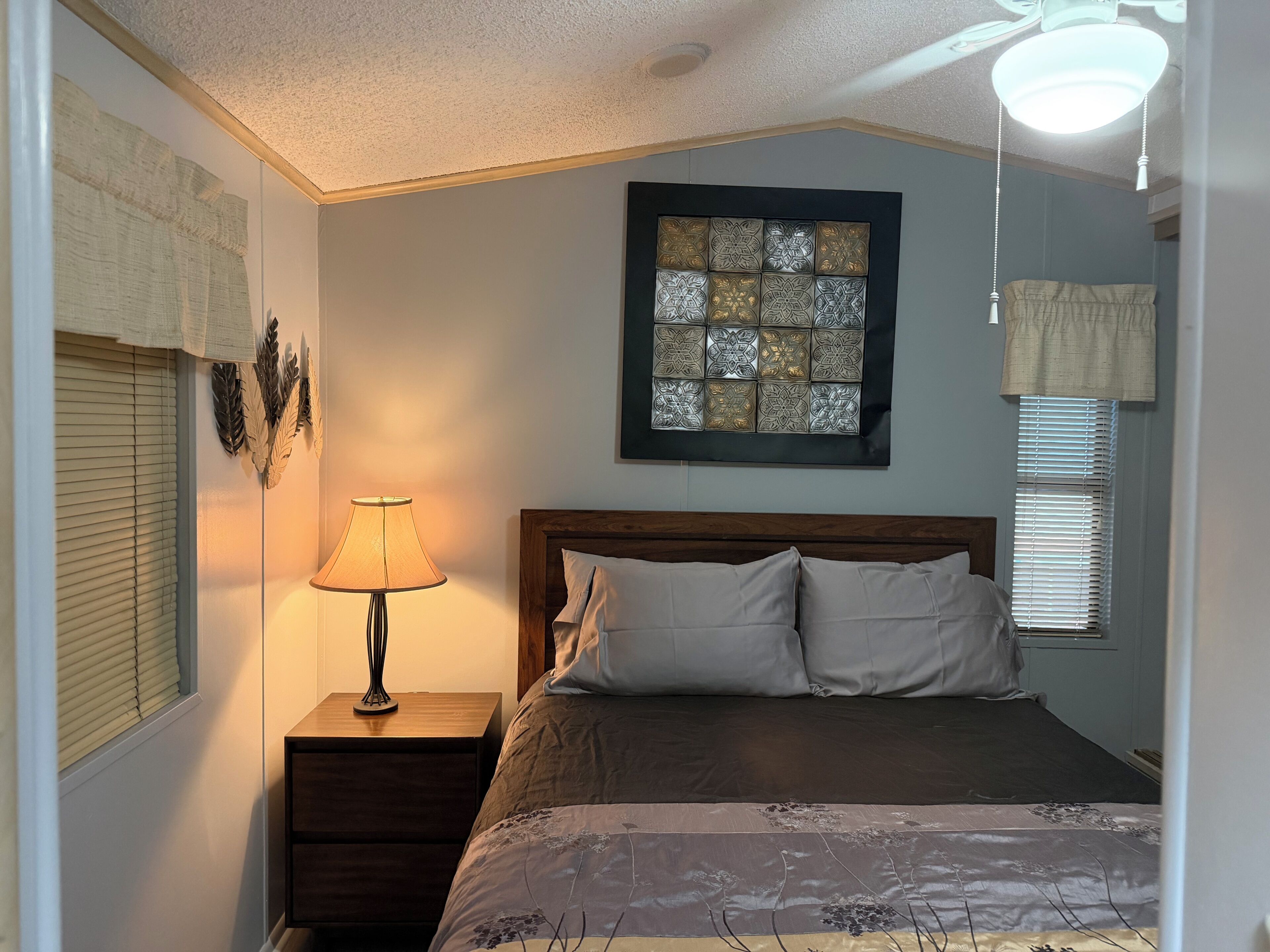 A bedroom with a bed, a nightstand, a lamp, a decorative wall piece, and a window with blinds.