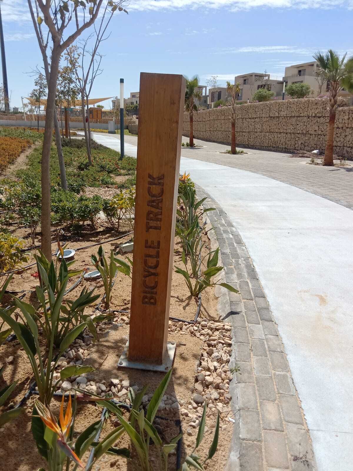 A wooden sign with 'BICYCLE TRACK' engraved, surrounded by plants and a paved path.