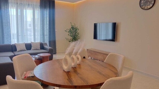 A modern living room with a round wooden dining table, white chairs, a grey sofa, a flat-screen TV, and a decorative plant. - Spacious 3-bedroom apartment in tranquil Cairo with AC (Cairo Governorate)