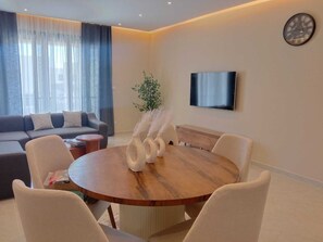 A modern living room with a round wooden dining table, white chairs, a grey sofa, a flat-screen TV, and a decorative plant. - Spacious 3-bedroom apartment in tranquil Cairo with AC (Cairo Governorate)