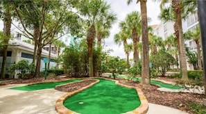 A landscaped area with palm trees, a green artificial turf putting green, and a building with a light-colored facade. - Two-Bedroom Suite at Calypso Cay Resort Florida (Kissimmee)