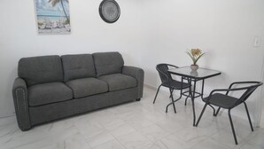Une pièce avec un canapé gris, une petite table ronde accompagnée de deux chaises, une horloge et un tableau représentant une scène de plage.