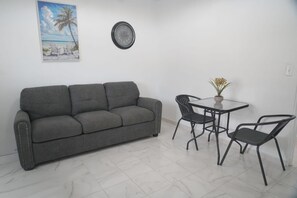 Ein Zimmer mit einem grauen Sofa, einem kleinen runden Tisch mit zwei Stühlen, einer Uhr und einem Bild mit einer Strandansicht.