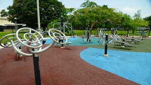 Un espace extérieur aménagé pour faire de l’exercice, équipé de divers appareils de musculation, notamment des appareils de type elliptique et des vélos d’appartement, et entouré d’un chemin pavé.