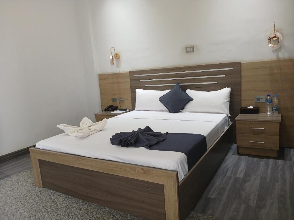 A hotel room with a wooden bed, white linens, a dark blue pillow, a bedside table with a lamp, and a nightstand with a phone and water bottles.