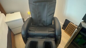 Een massagestoel in een kamer met een bed, een klein tafeltje en een kast.