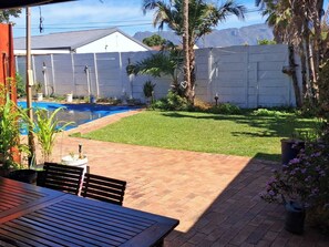 A backyard with a swimming pool, a patio table, and a fenced area with grass and plants. - Adventure Cape Town 1 (Cape Town)